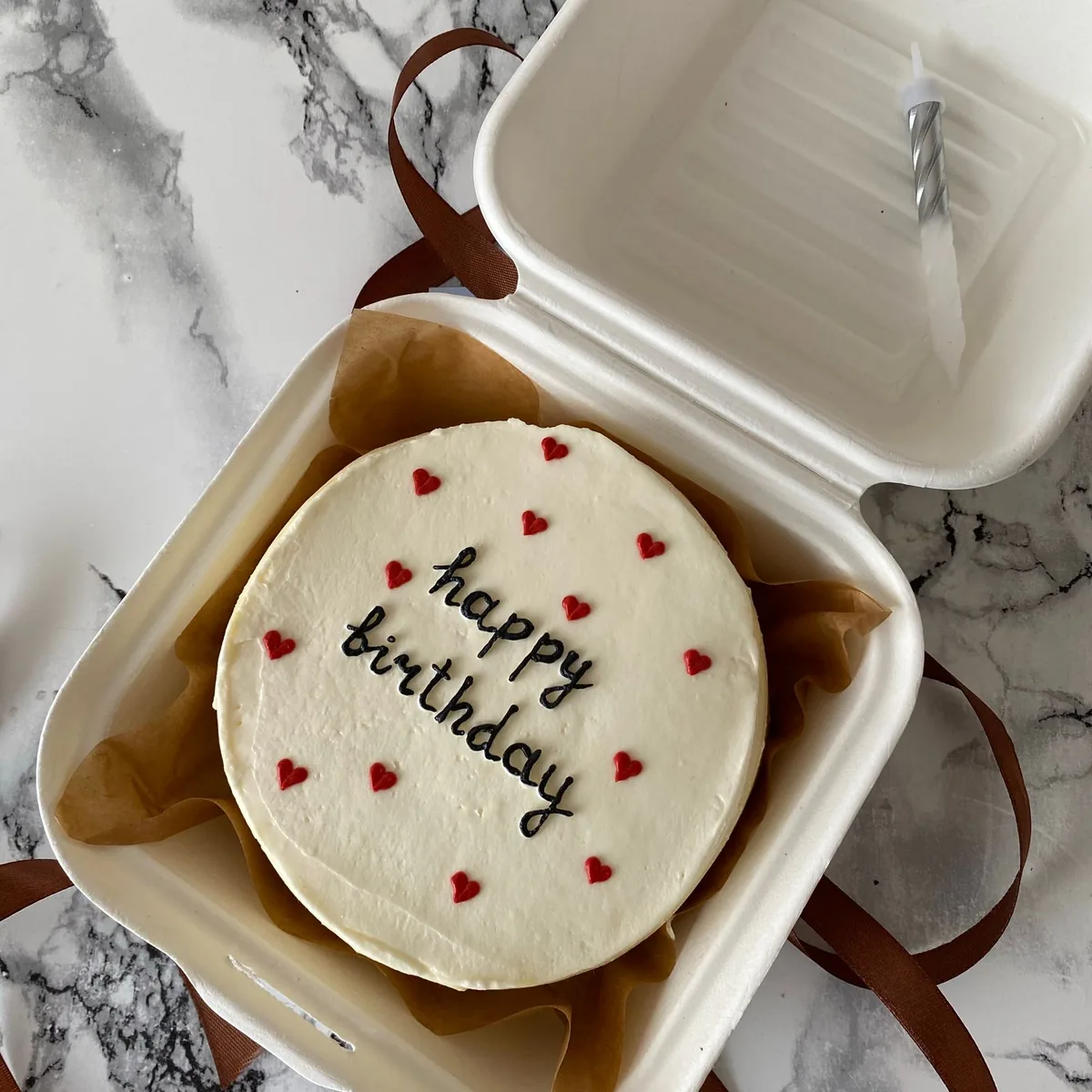 White paperboard clamshell bento cake box holding a small birthday cake with heart sprinkles